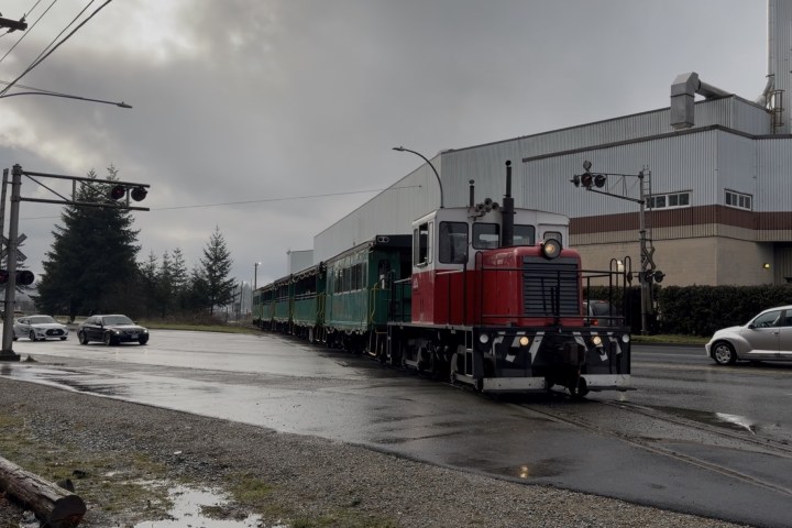 Small red train crossing a road in an industrial area on an overcast day.